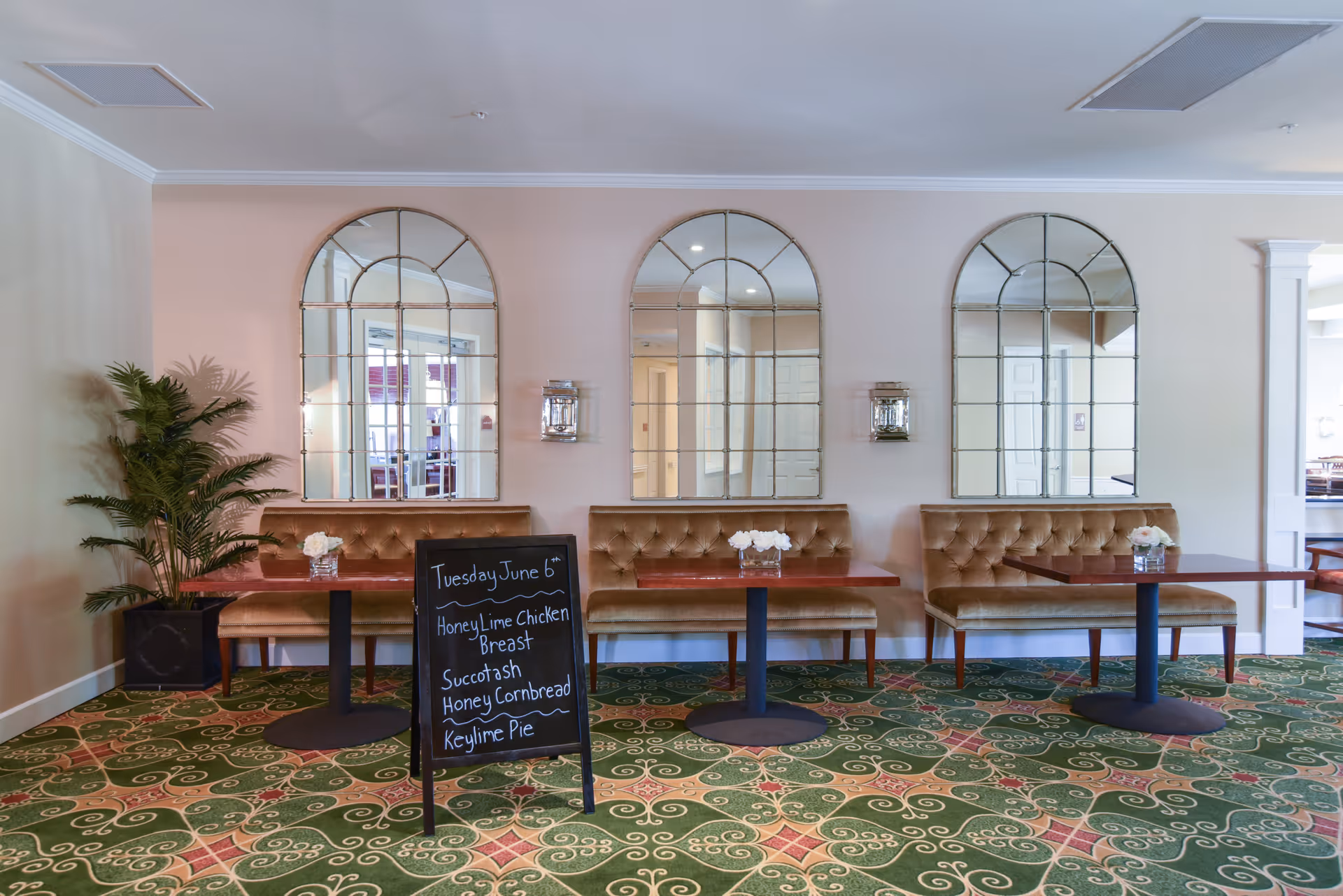 Dining area with three wooden tables and cushioned bench seating against a wall with three large arched mirrors. A chalkboard sign displays the menu for Tuesday, June 6th, featuring honey lime chicken breast, succotash, honey cornbread, and key lime pie. A potted plant is placed on the left side, and the floor is covered with a green patterned carpet.