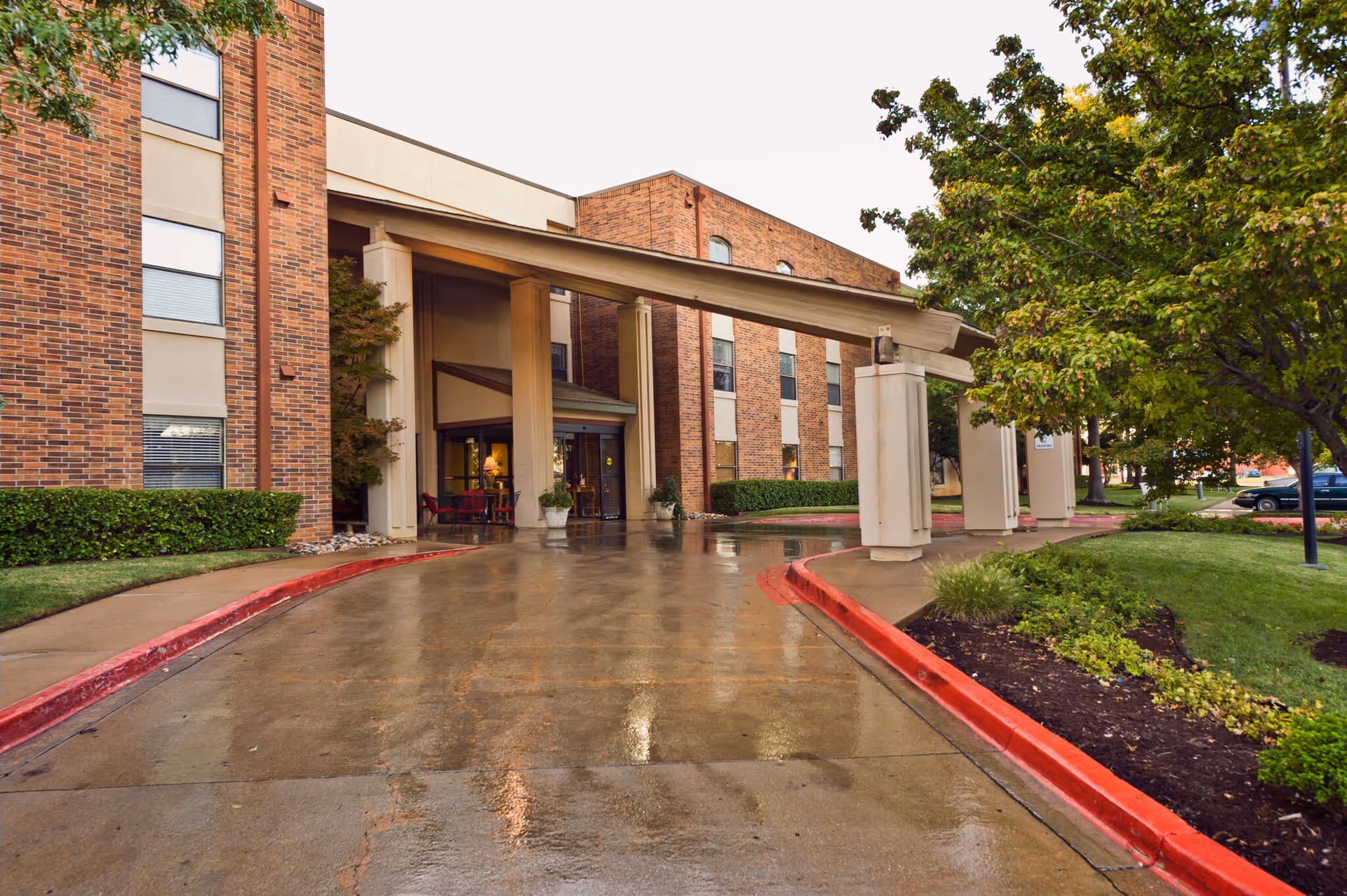 Entrance of a brick senior living facility with a covered drop-off area supported by large columns. The driveway is wet, reflecting the building and surrounding greenery, with neatly trimmed bushes and trees on either side.