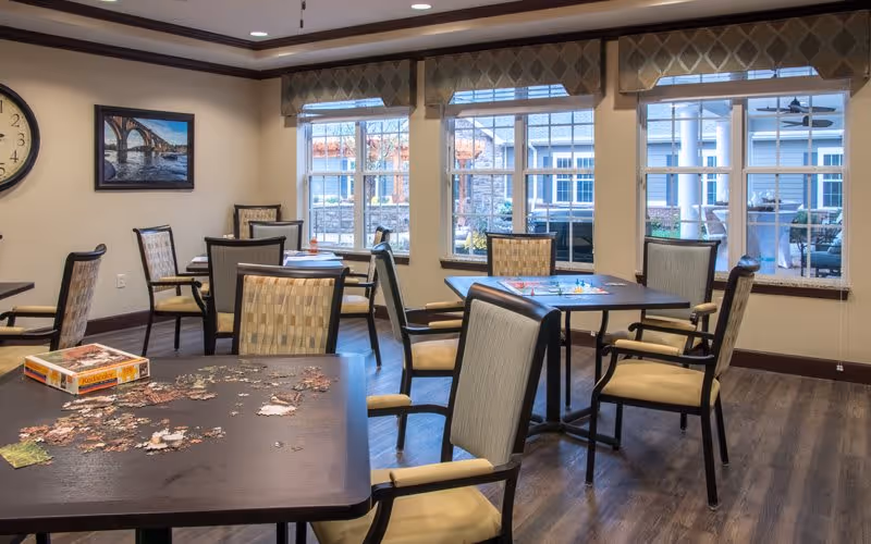 A well-lit common room with multiple tables and chairs arranged for group activities. One table has a partially completed jigsaw puzzle and a puzzle box, while another table has a board game set up. Large windows with valances allow natural light to fill the room, and a clock and framed picture hang on the wall.