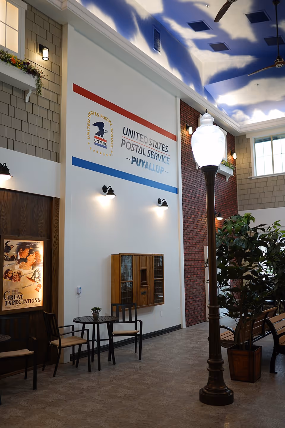 Indoor communal area styled like a small-town street with a 'United States Postal Service – Puyallup' wall sign, a tall decorative lamppost, seating, and plants.
