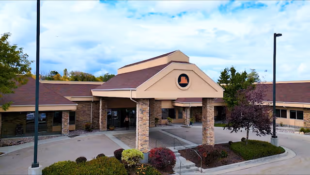 Exterior view of Monument Health Taylorsville facility showing a single-story building with a covered entrance supported by stone pillars, surrounded by landscaped bushes and trees under a partly cloudy sky.