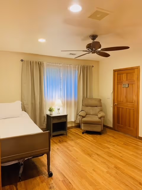 A cozy room with a single bed on the left, a small nightstand with a lamp and a small plant in the center, a comfortable recliner chair near a window with curtains, and a wooden door labeled 'Rest Room' on the right. The room has wooden flooring and a ceiling fan with lights.