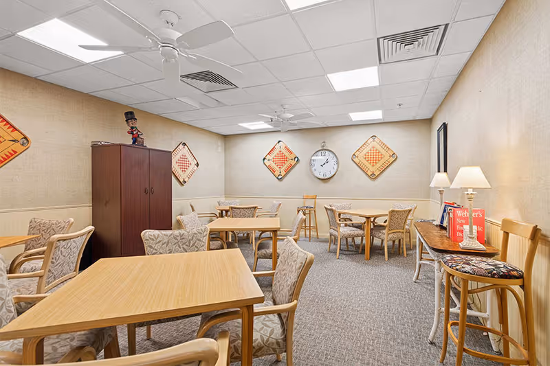 A cozy room with several wooden tables and cushioned chairs arranged for group seating. The walls are decorated with large game boards and a clock. There is a wooden cabinet in the corner and a side table with two lamps and books. The ceiling has recessed lighting and ceiling fans.