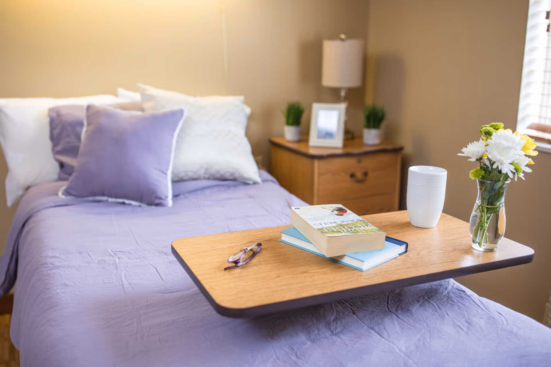 A neatly made bed with lavender bedding and multiple pillows in a softly lit room. A wooden overbed table holds a pair of glasses, two books, a white cup, and a small vase with white and yellow flowers. In the background, there is a wooden nightstand with a lamp, two small potted plants, and a framed photo.
