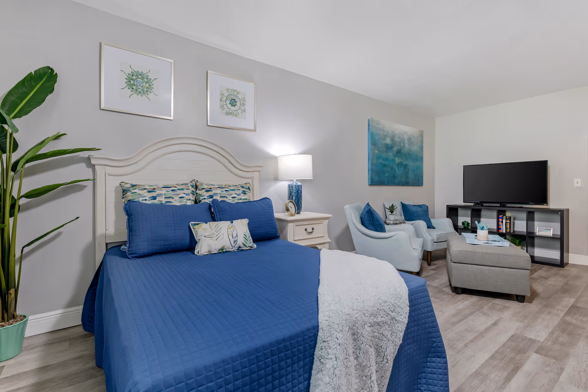 A cozy bedroom with a white wooden headboard bed dressed in blue bedding and decorative pillows. There is a white nightstand with a blue lamp and a clock. Two framed botanical prints hang above the bed. To the right, there are two light blue armchairs with blue cushions, a gray ottoman, and a black TV stand with a flat-screen TV and some books. A large green potted plant is on the left side of the bed. The room has light gray walls and wood flooring.