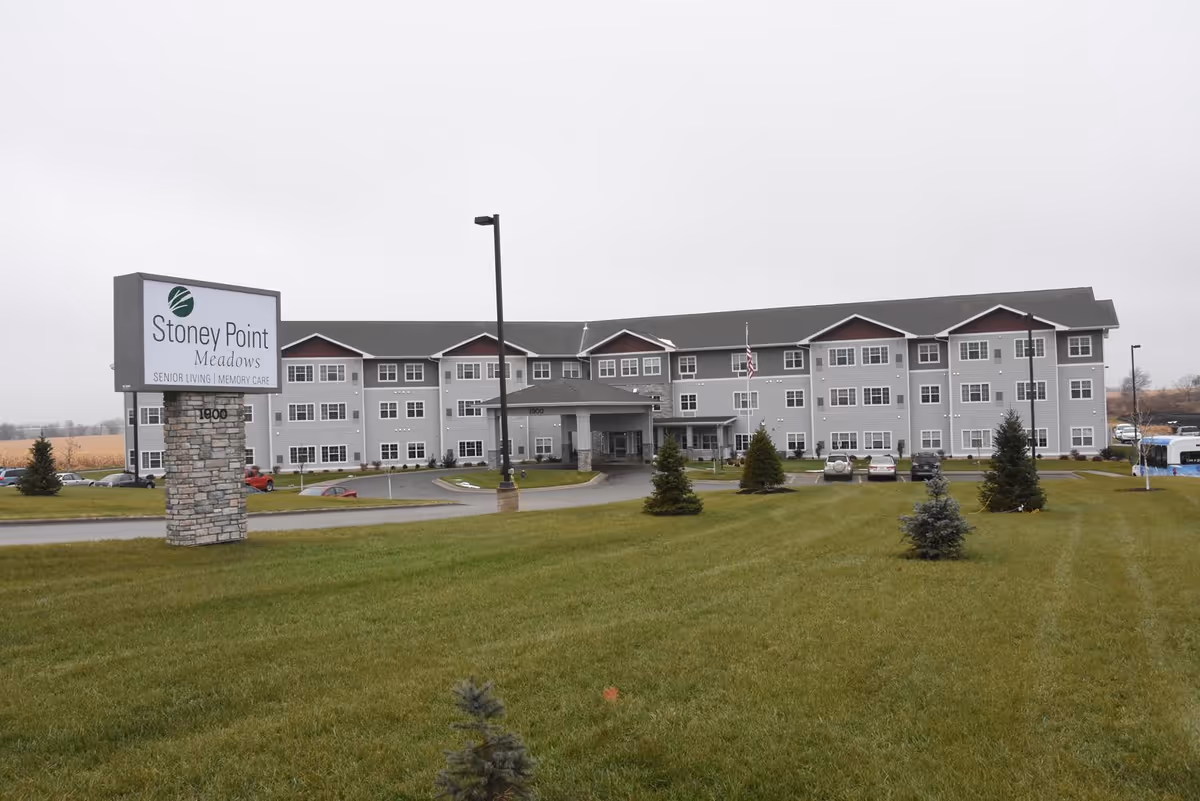 Exterior view of Stoney Point Meadows, a senior living and memory care facility. The building is a large, three-story structure with gray siding and white trim. There is a sign in the foreground on a stone base displaying the facility's name. The lawn is green with small trees planted, and several cars are parked near the entrance.