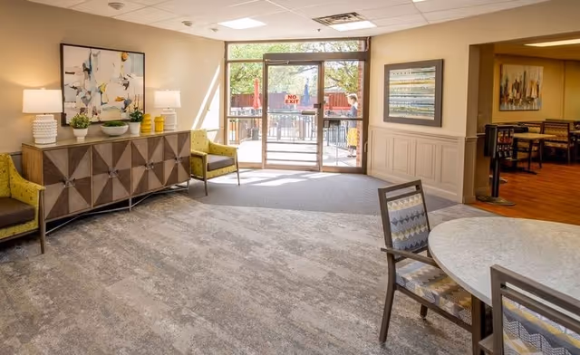 Bright communal lounge with chairs, a sideboard, a round table and glass doors leading to an outdoor patio.
