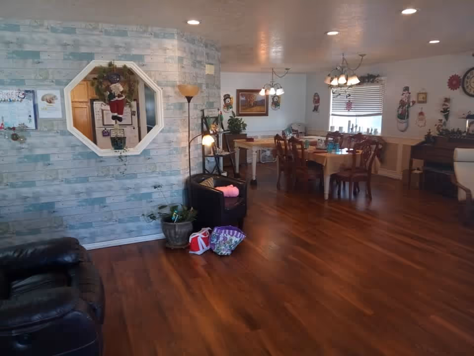 Interior view of a senior living facility dining and lounge area with wooden flooring, a dining table with chairs, a piano, wall decorations including Christmas-themed items, a floor lamp, and a small seating area with a black armchair and plants.
