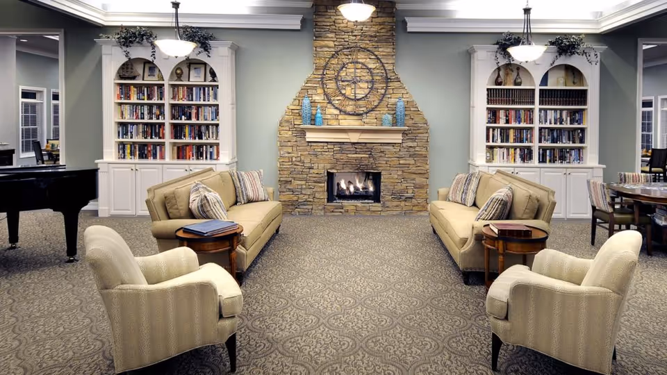 A cozy senior living facility common area with two beige sofas facing each other, each with striped throw pillows. Two beige armchairs are positioned in front of the sofas. A stone fireplace with a large decorative clock above it is centered on the back wall. On either side of the fireplace are white built-in bookshelves filled with books and decorative items. The room has patterned carpet flooring and soft lighting from ceiling fixtures. A black grand piano is visible on the left side, and a dining area with tables and chairs is partially visible on the right.