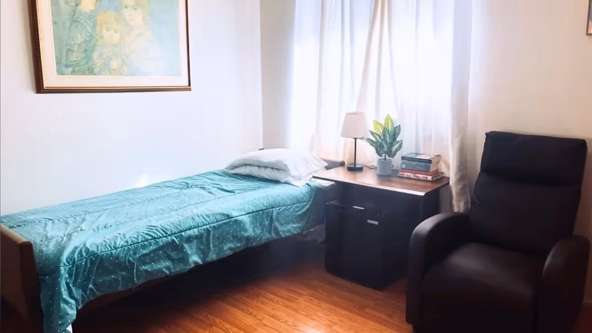 A small bedroom with a single bed covered in a teal blanket with white polka dots, a white pillow, a wooden nightstand with a lamp, a potted plant, and a stack of books. Next to the nightstand is a black recliner chair. A framed picture hangs on the white wall above the bed, and sheer white curtains cover the window behind the nightstand.