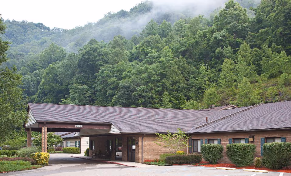 Front entrance of a single-story brick senior living building with a covered drop-off and wooded hills behind it.