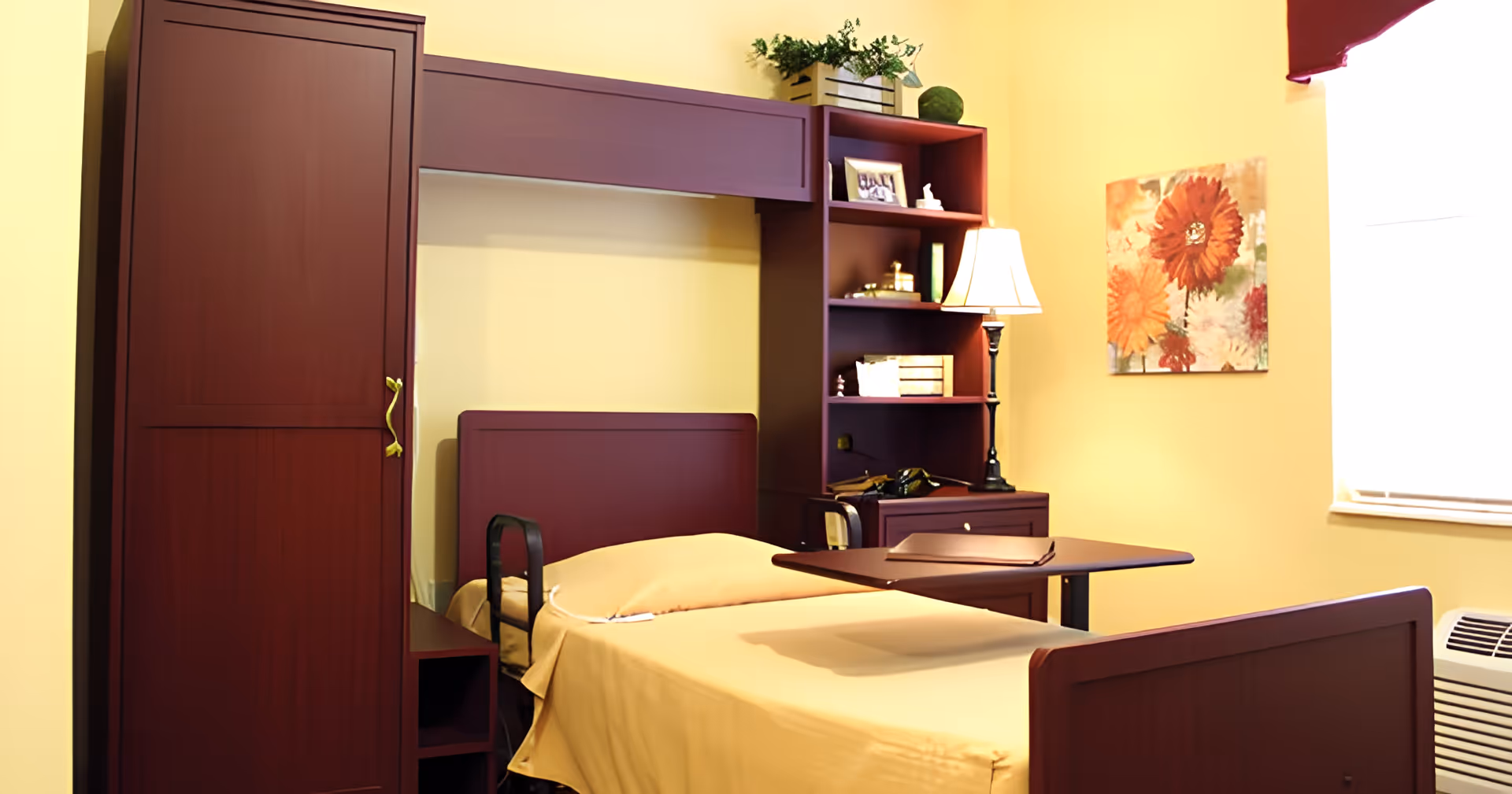 A senior living facility bedroom with a single bed covered in a beige blanket, a maroon wooden headboard and matching furniture including a wardrobe, shelves, and a bedside table with a lamp. There is a floral painting on the yellow wall and a window with a white blind letting in natural light.