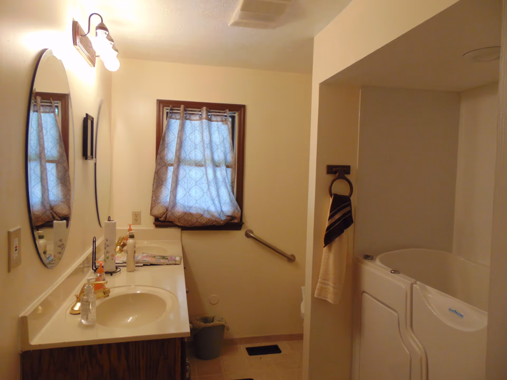 A bathroom with a single sink vanity featuring a round mirror above it, a window with patterned curtains, a grab bar on the wall, a towel hanging on a ring, and a walk-in bathtub with a door.