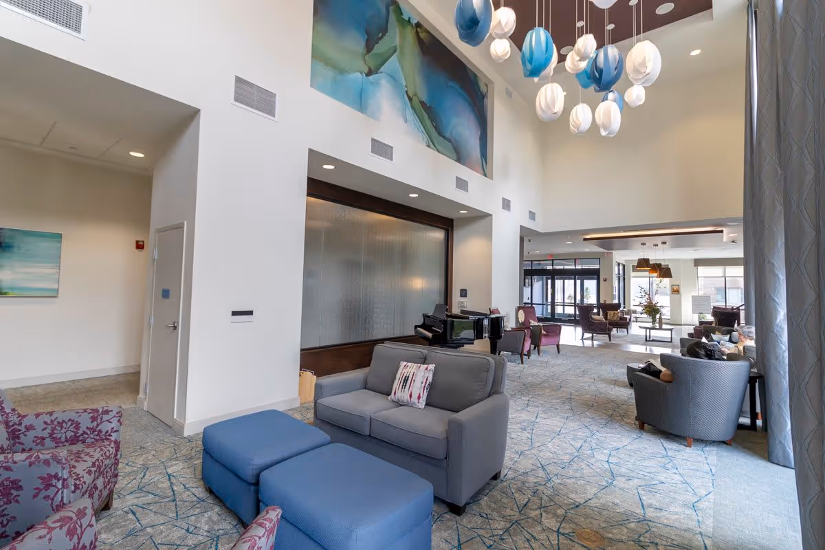 A spacious and well-lit living room area in a senior living facility with modern furniture including gray sofas, blue ottomans, and purple armchairs. The room features a grand piano, large abstract artwork on the wall, and a contemporary chandelier with blue and white hanging lights. Large windows and glass doors allow natural light to fill the space.