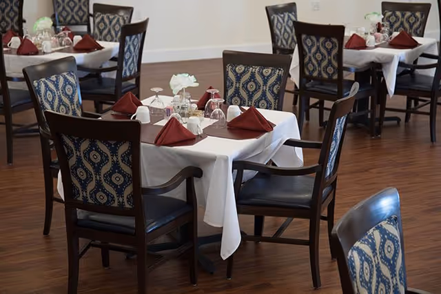 Dining room with several tables set with white tablecloths, folded burgundy napkins, glassware, and patterned chairs.