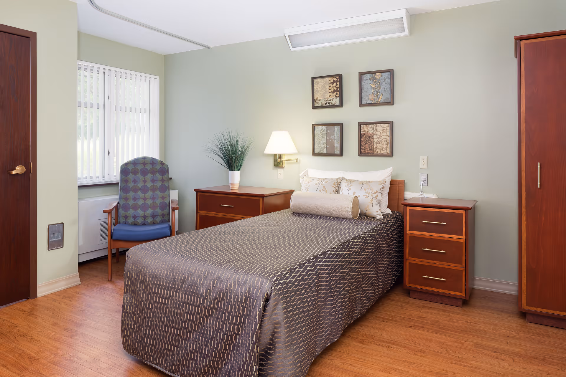 A neatly arranged bedroom in a senior living facility featuring a single bed with a patterned bedspread, two floral pillows, and a cylindrical bolster pillow. On either side of the bed are wooden nightstands with drawers, one holding a potted plant and the other a wall-mounted lamp. A patterned armchair is positioned near a window with vertical blinds, and a wooden wardrobe stands to the right. The walls are painted light green and decorated with four framed floral artworks above the bed.