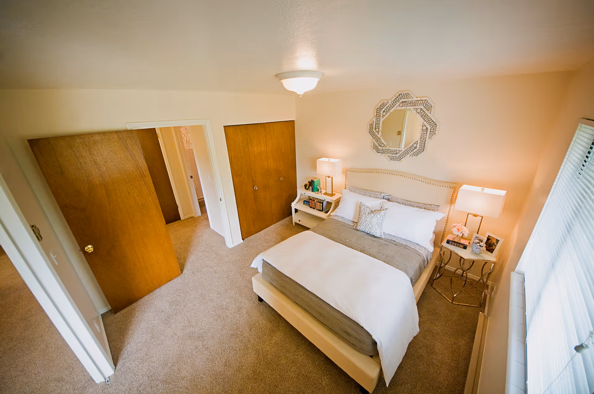 A cozy bedroom with a neatly made bed featuring white and gray bedding. There are two bedside tables with lamps, books, and decorative items. A decorative mirror hangs above the bed. The room has beige carpet, wooden closet doors, and a window with blinds letting in natural light.
