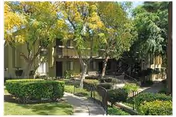Outdoor view of a senior living facility with a pathway leading through a landscaped garden area featuring green bushes and trees. The building has balconies and staircases visible, surrounded by greenery under a clear sky.