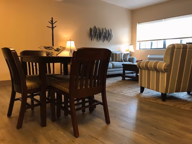 A cozy living area with a wooden dining table and four chairs in the foreground. In the background, there is a light-colored sofa with cushions, a striped armchair, two table lamps, a decorative wall piece, and a large window letting in natural light.