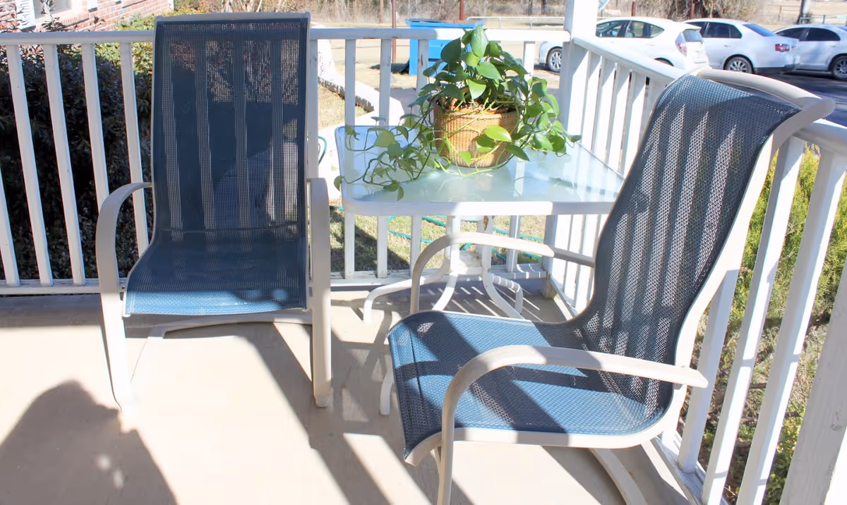 Two blue mesh patio chairs and a small white table with a potted green plant on a porch with white railing. Cars and greenery are visible in the background.