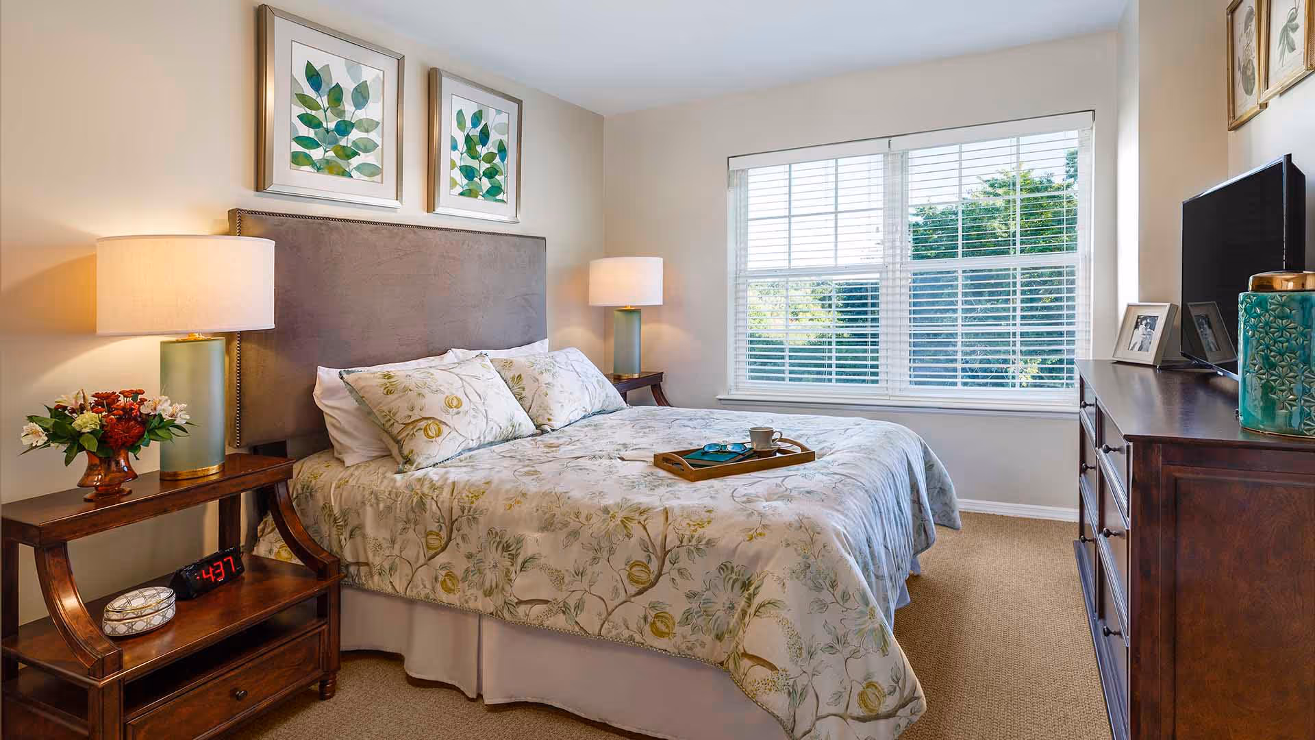 Bright, neatly decorated bedroom featuring a bed with floral bedding, two nightstands with lamps, a dresser with a TV, and a large window with blinds.