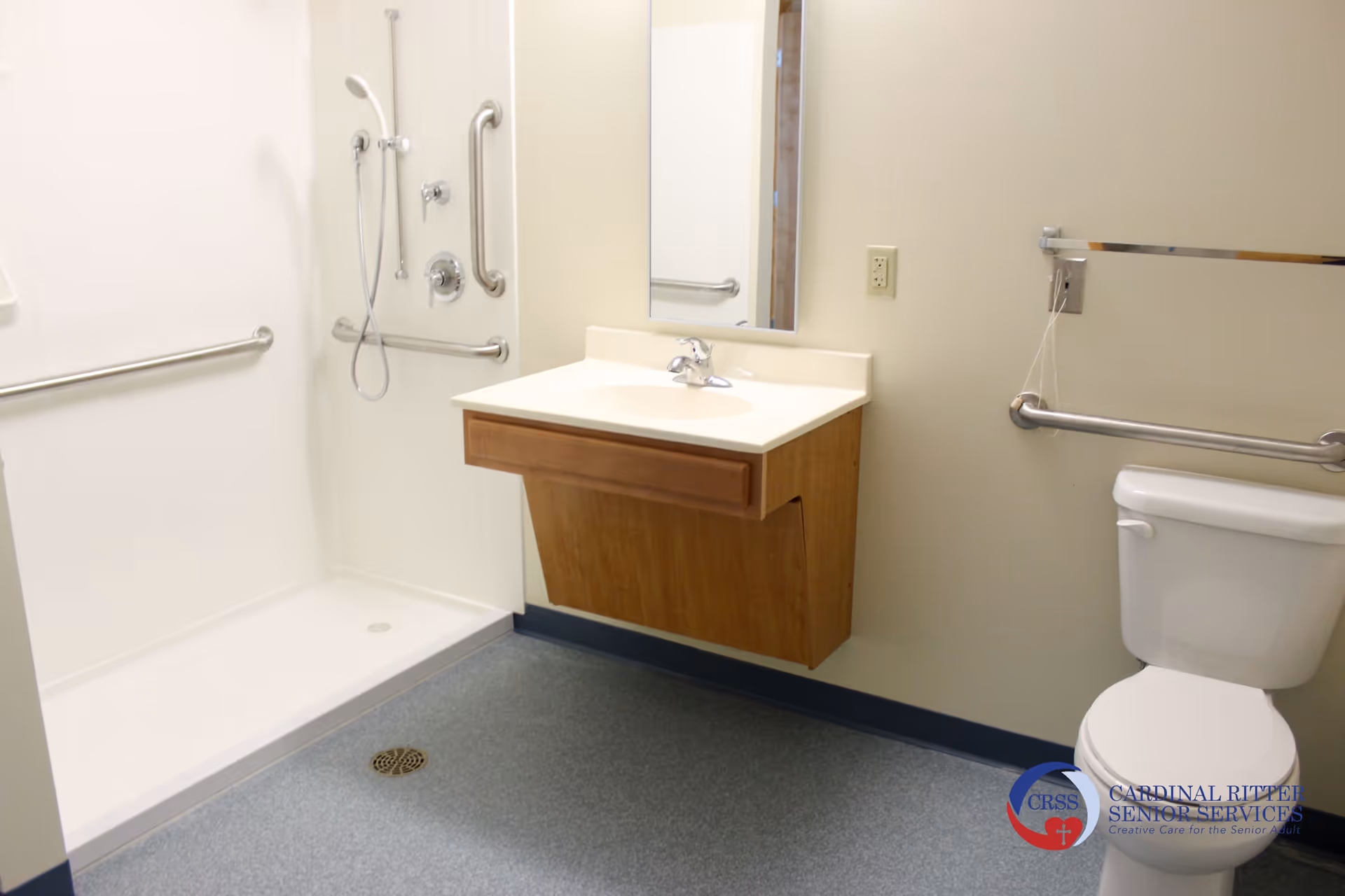 A clean, accessible bathroom featuring a walk-in shower with grab bars, a wall-mounted sink with a mirror above it, and a toilet with a grab bar beside it. The floor is covered with a non-slip material, and the walls are light-colored.