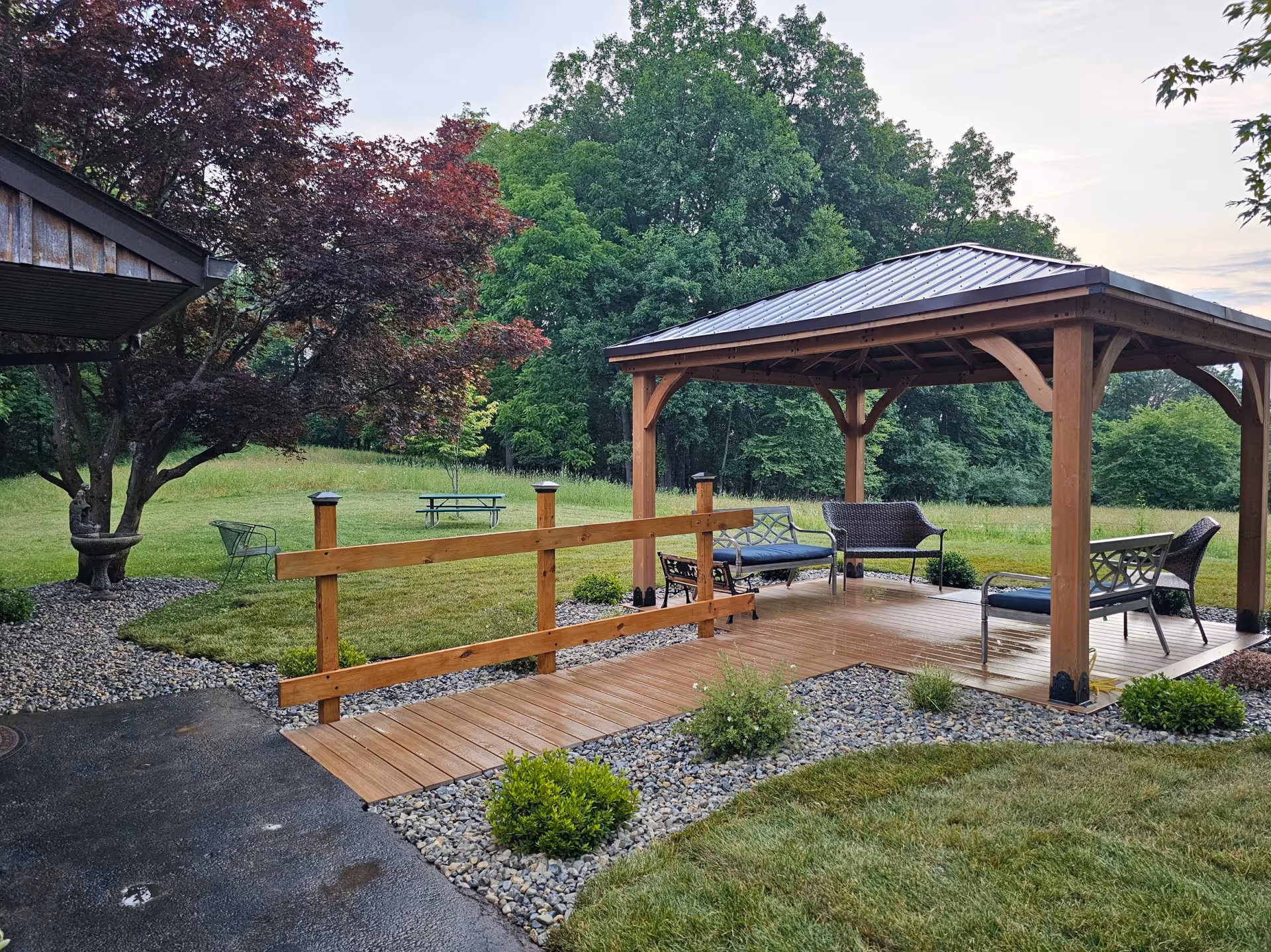 Outdoor wooden gazebo with seating, a wooden walkway, and manicured lawn surrounded by trees.