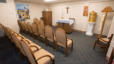 A small chapel room with two rows of beige cushioned chairs facing an altar covered with a white cloth. The altar has two candles and a crucifix mounted on the wall behind it. Religious artwork and statues are displayed around the room, including a painting on the left wall and a golden tabernacle on a pedestal to the right.