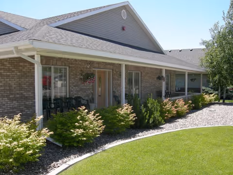 Exterior view of a single-story brick building with a covered porch, several windows, hanging flower pots, and green shrubs along the front. The lawn is well-maintained with a curved edge separating the grass from a rock garden. Trees are visible in the background under a clear blue sky.
