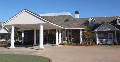 Front exterior view of Arbor Senior Apartments building with a covered entrance supported by white columns, stone and siding facade, and a driveway in front.