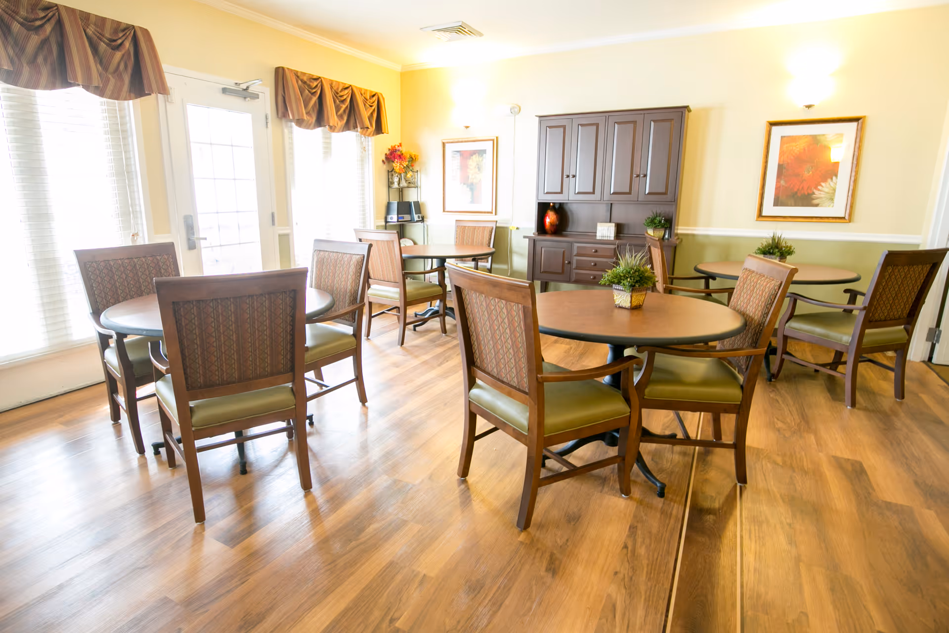 A bright and clean dining area with several round wooden tables and cushioned chairs arranged neatly on a wooden floor. The room has large windows with blinds and valances, a door leading outside, a wooden cabinet with decorative items, framed floral artwork on the walls, and small potted plants on the tables.