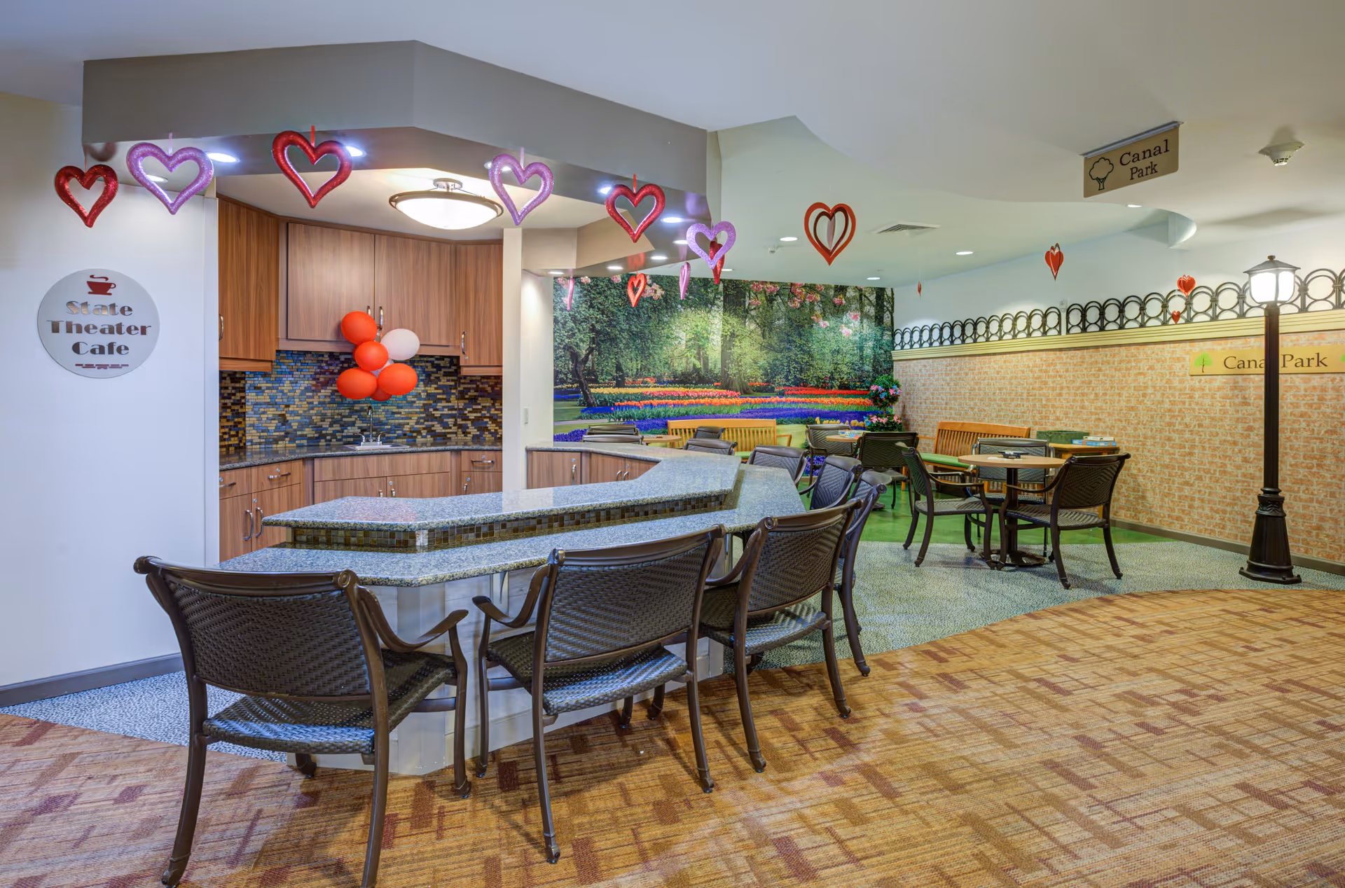 Indoor cafe area with a curved granite countertop and several chairs. The back wall features a kitchen area with wooden cabinets and a mosaic tile backsplash. Red and white balloons are attached to the cabinets. Heart-shaped decorations hang from the ceiling. The far wall has a large mural of a colorful garden with trees and flowers. Several tables and chairs are arranged in the seating area. A sign on the left reads 'State Theater Cafe' and signs on the right indicate 'Canal Park'.