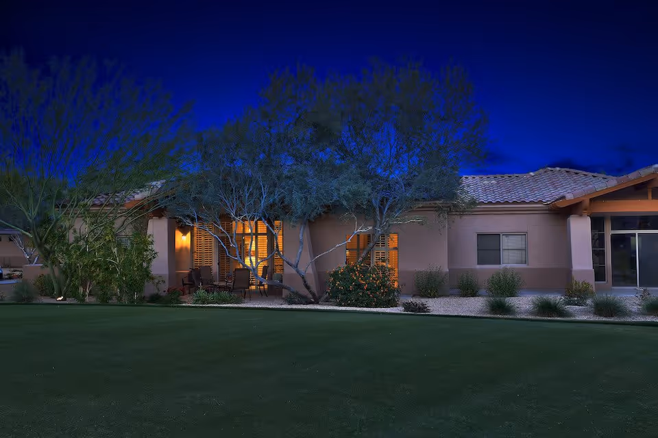 Exterior view of a single-story building at night with warm lights glowing from inside. The building has a tiled roof and is surrounded by trees, bushes, and a well-maintained lawn in the foreground.