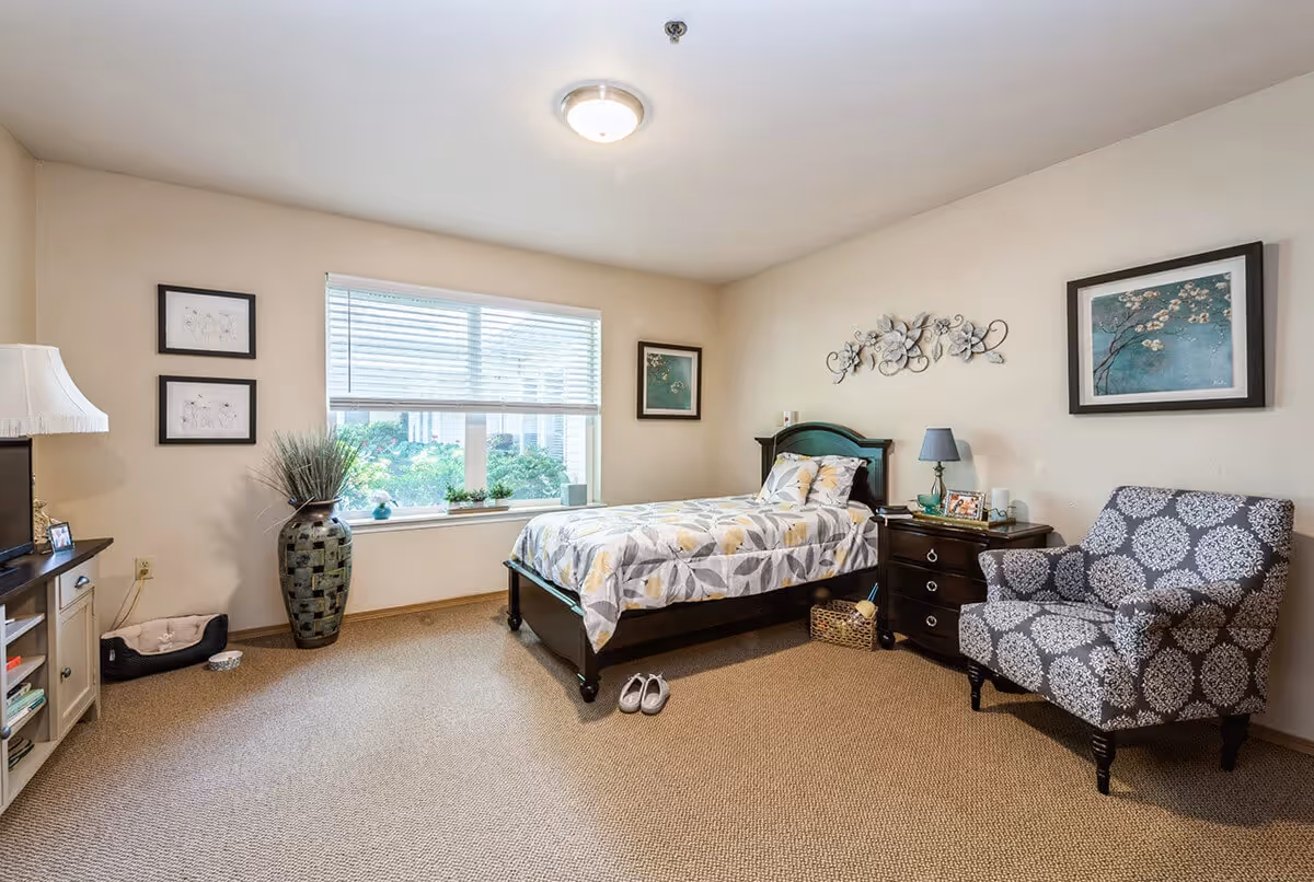 A cozy bedroom with a single bed covered in a floral patterned comforter, a dark wooden nightstand with a lamp and framed photos, a patterned armchair, a large window with blinds letting in natural light, a tall decorative vase with plants, and framed artwork on the walls.