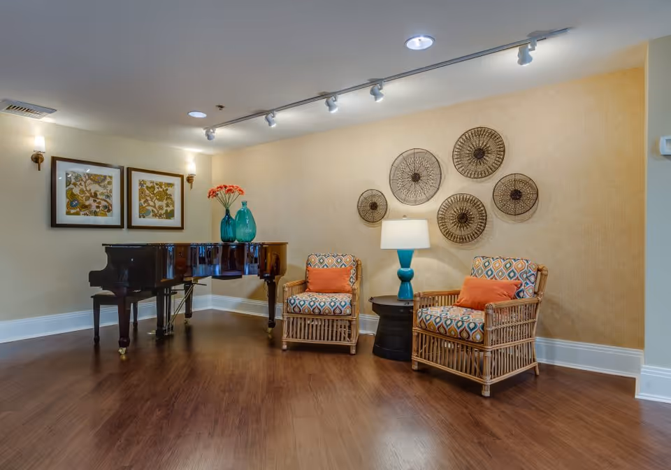 A cozy interior seating area with two patterned armchairs featuring orange cushions, a small round black side table with a blue lamp, and a grand piano adorned with two blue vases and orange flowers. The walls are decorated with framed artwork and circular metal wall hangings. The floor is wooden, and the room is softly lit with wall sconces and ceiling track lights.