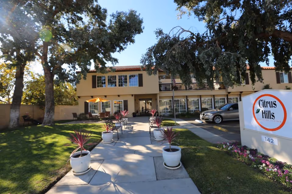 Exterior view of Citrus Hills Senior Living facility showing a two-story building with large windows, a walkway lined with potted plants, outdoor seating with an umbrella, trees, a parked car, and a sign with the facility name and address number 142.