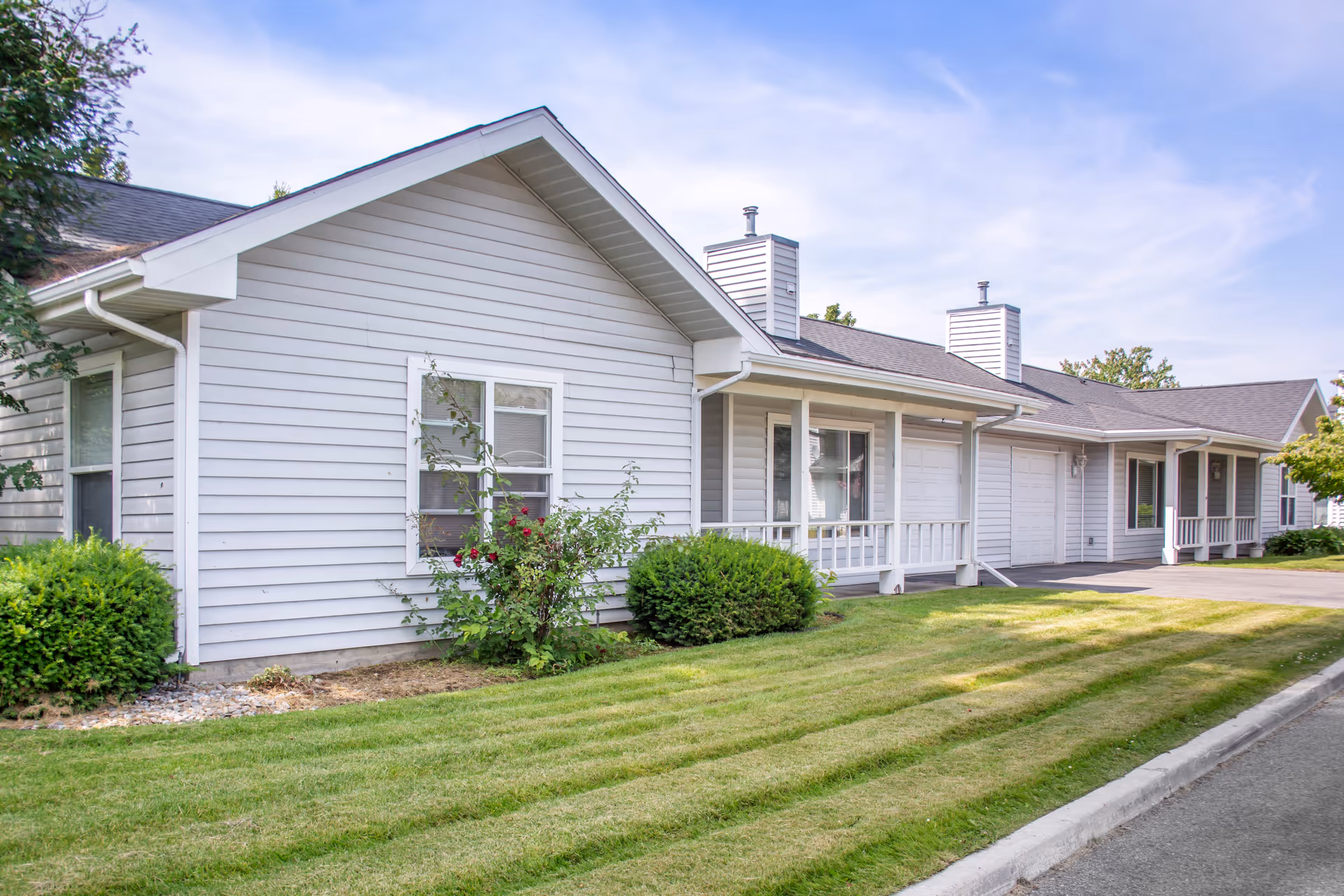 Exterior view of a single-story residential building with white siding, a small porch, and attached garages. The building is surrounded by neatly trimmed grass, bushes, and a few small trees under a partly cloudy sky.
