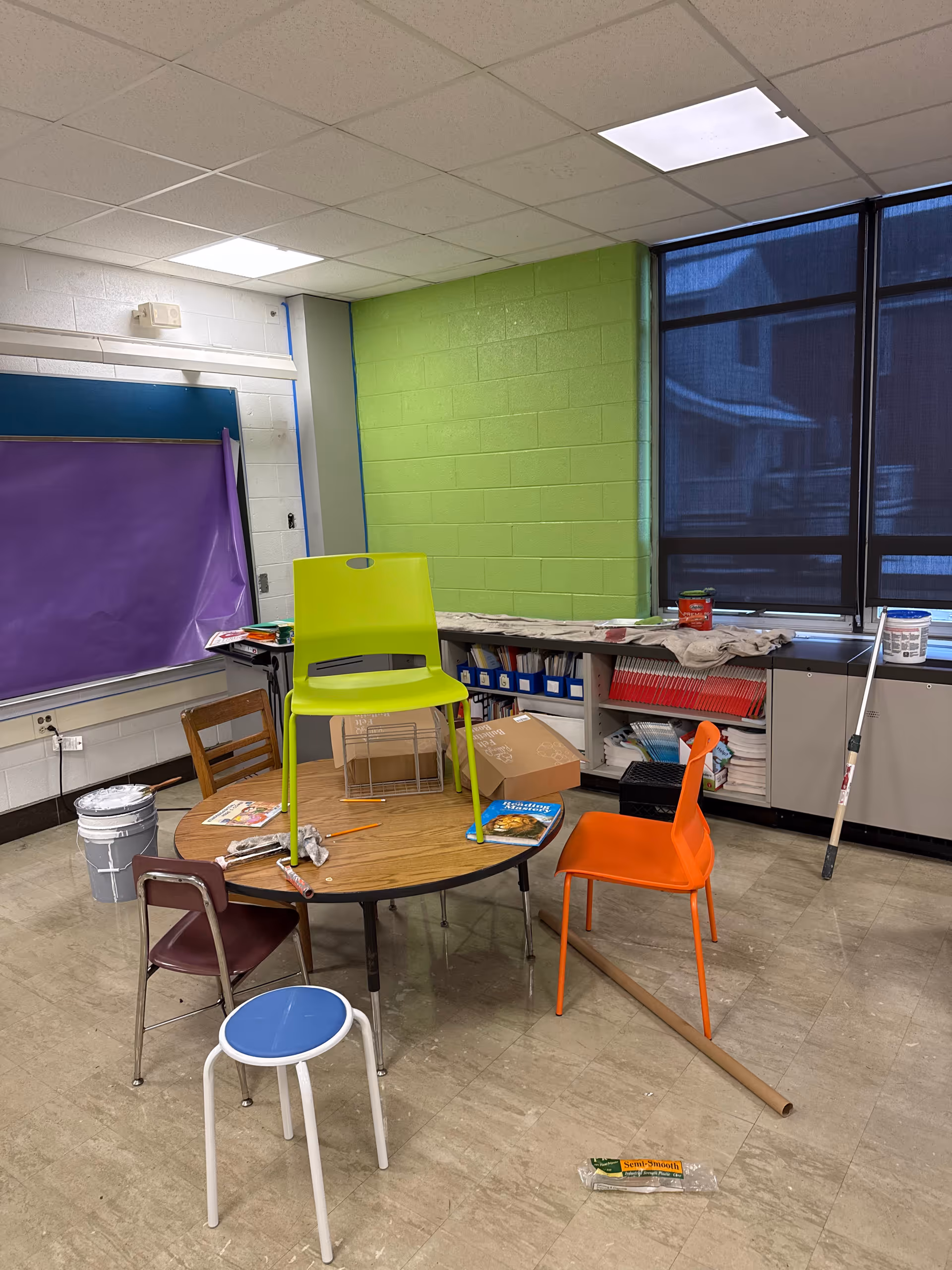 Colorful classroom-like activity room with stacked chairs, round table, and construction clutter.