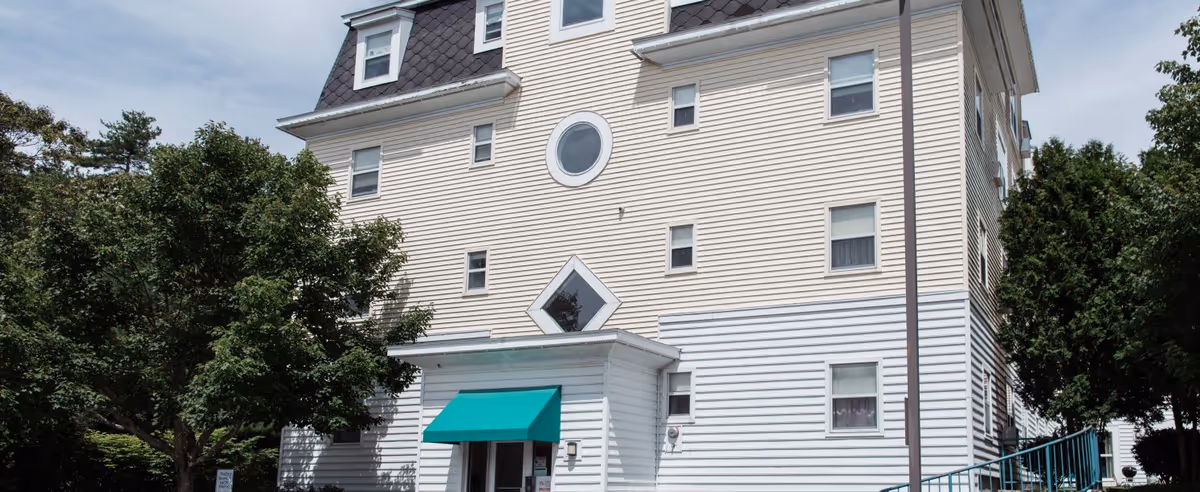 A multi-story white senior living building with a green awning over the entrance, set among trees.