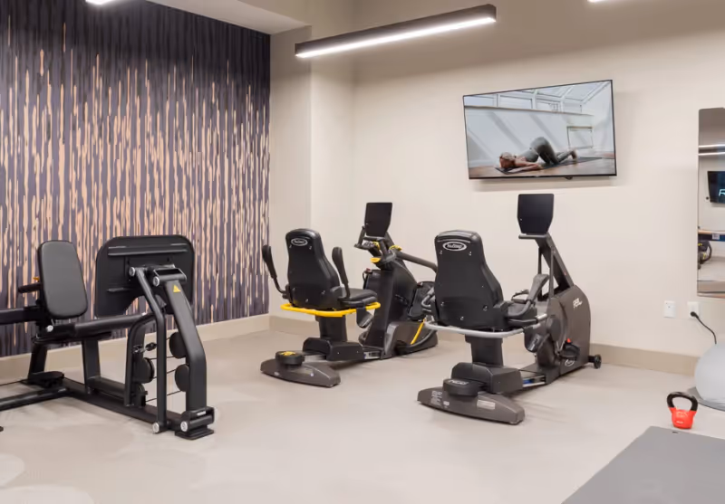 A fitness room with exercise equipment including two recumbent stationary bikes and a leg press machine. A large flat-screen TV mounted on the wall displays a person doing a floor exercise. The room has a patterned accent wall and neutral-colored flooring.