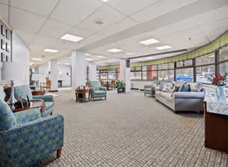 Spacious senior living common lounge with sofas and armchairs arranged near large windows and overhead lighting.