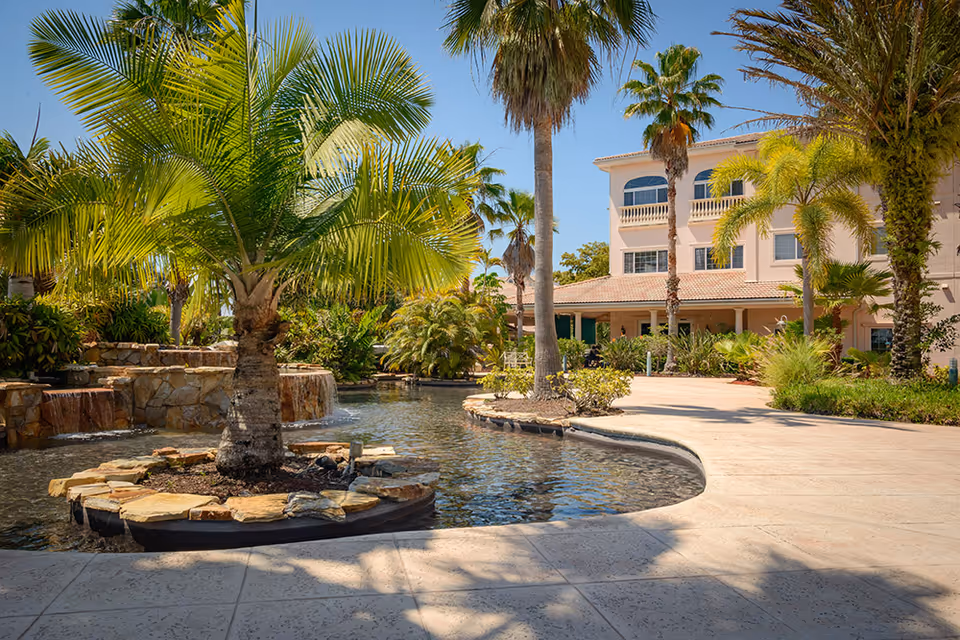 A landscaped courtyard with a small pond, palm trees, and the front of a multi-story building.