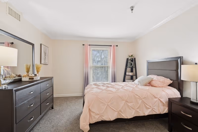 A bedroom with a large window covered by pink and gray ombre curtains. The bed has a pink comforter and pillows, and there is a dark wooden headboard. To the right of the bed is a nightstand with a lamp. On the left side of the room is a dark wooden dresser with a large mirror above it and decorative items on top. The walls are light beige and the floor is carpeted in gray.