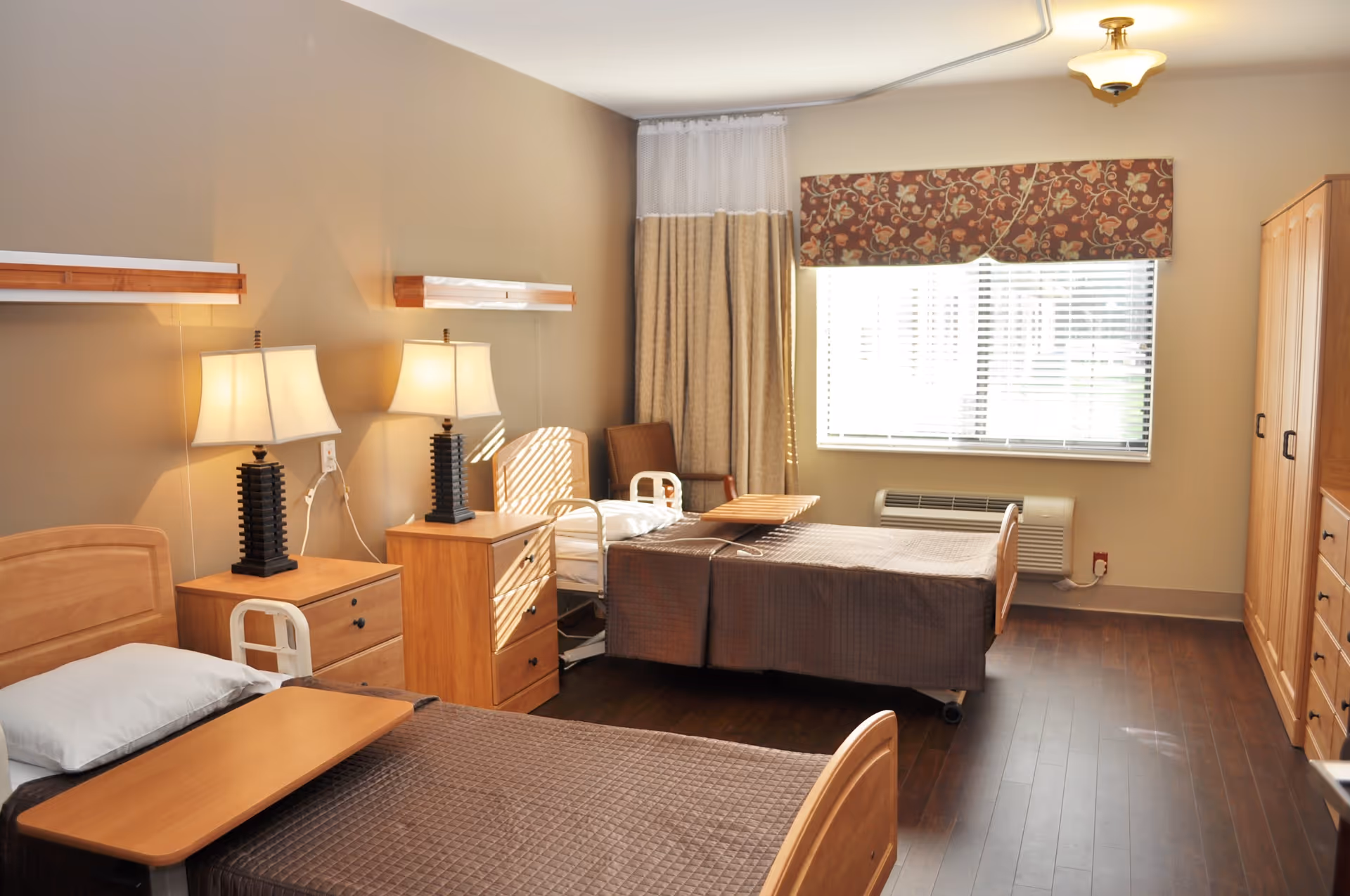 A well-lit room with two single beds, each with a brown quilted bedspread and a wooden overbed table. Between the beds are two wooden nightstands with matching lamps. A window with blinds and a floral valance lets in natural light. There is a wooden wardrobe and a chair in the corner, and the floor is dark wood.