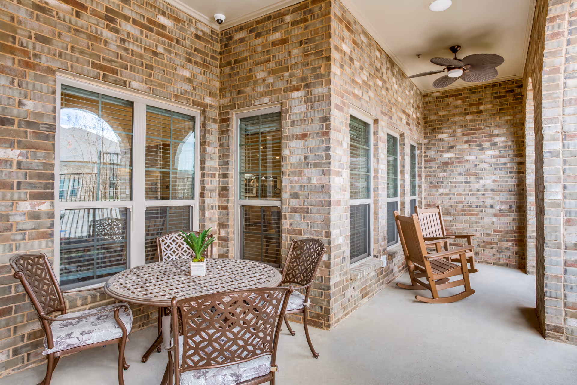 Covered brick patio with a round metal table and chairs and wooden rocking chairs along windowed wall.