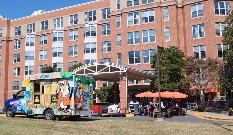 Exterior view of a multi-story brick assisted living facility with a covered entrance. A colorful Kona Ice truck is parked on the grass near the entrance. Several people are seated at tables with orange umbrellas outside the building, enjoying the sunny day.