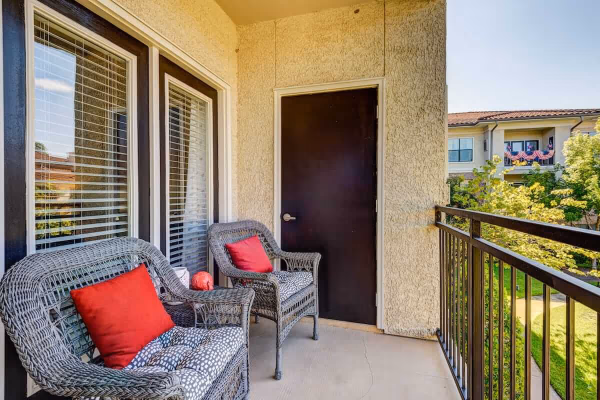 A small outdoor balcony with two wicker chairs featuring red cushions and patterned seat cushions. The balcony has a black metal railing and overlooks a green garden area with trees and a neighboring building decorated with patriotic bunting.