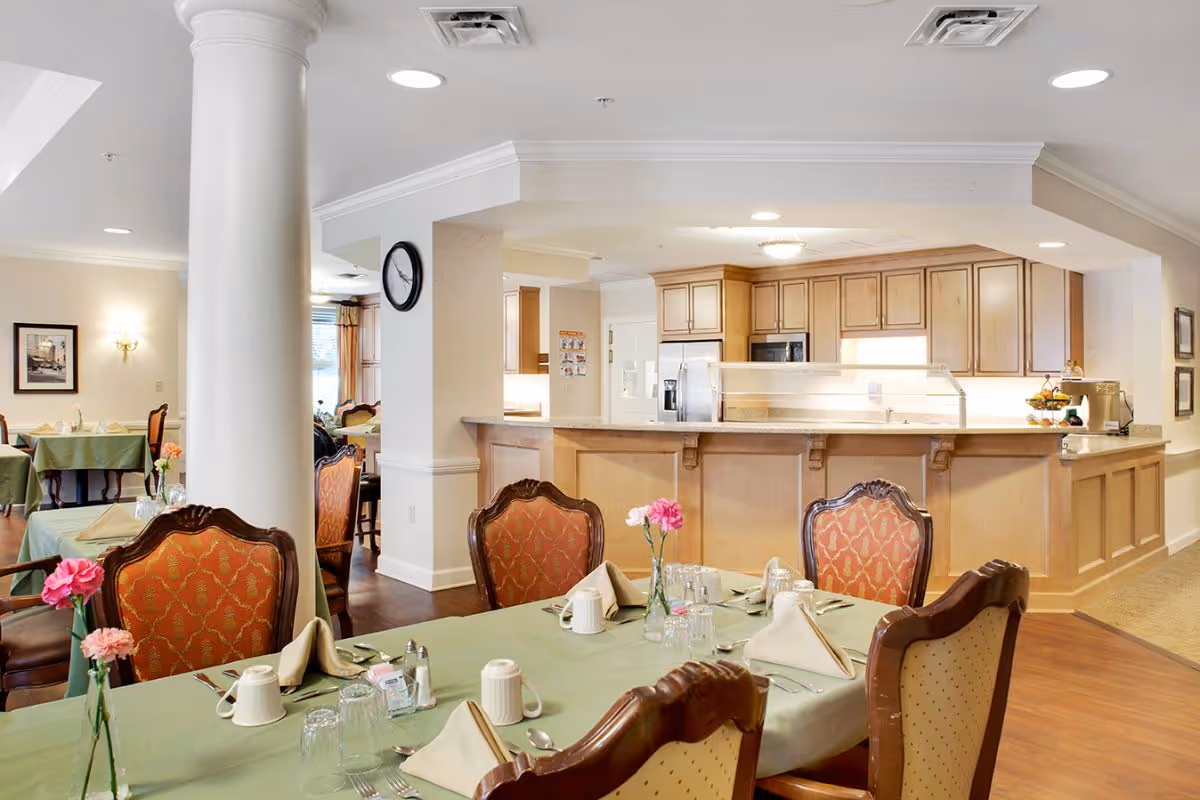 Dining room with set tables, upholstered chairs, flowers on the tables, and a serving counter/open kitchen in the background.