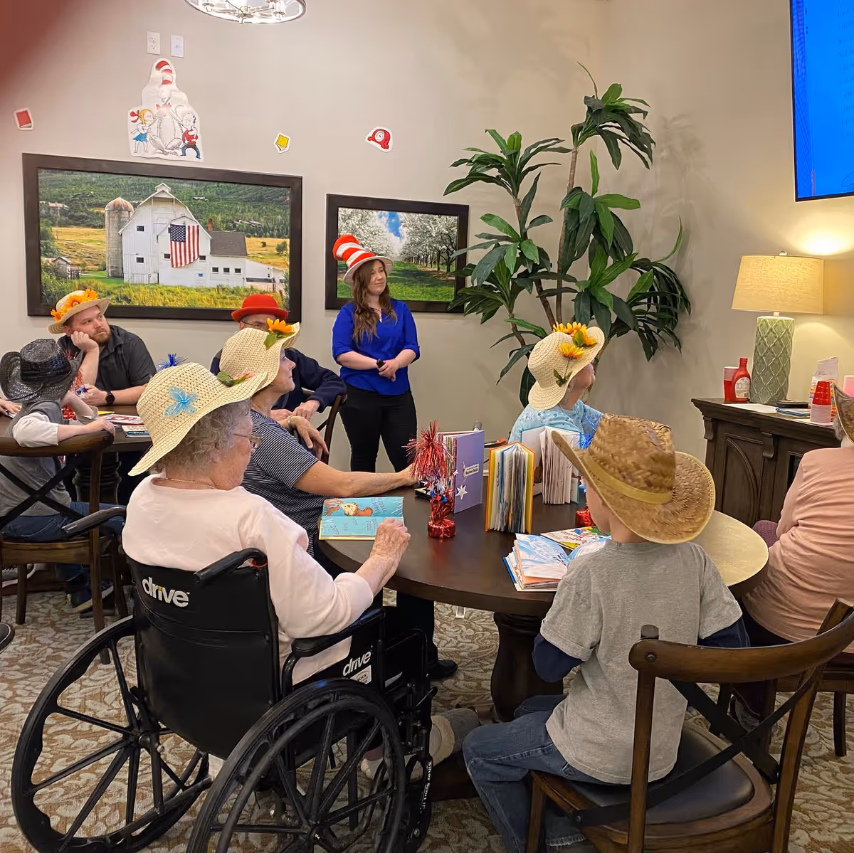 A group of elderly people and a child wearing various hats sit around a table in a common room. A woman in a blue shirt and a red and white striped hat stands near the wall, engaging with the group. The room has framed pictures on the wall, a large plant, and a lamp on a side table.