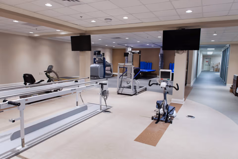 Interior view of a rehabilitation or physical therapy room with exercise equipment including parallel bars, stationary bikes, and other therapy machines. The room has beige walls, a light-colored floor, and two large flat-screen TVs mounted on the ceiling. A hallway is visible in the background.